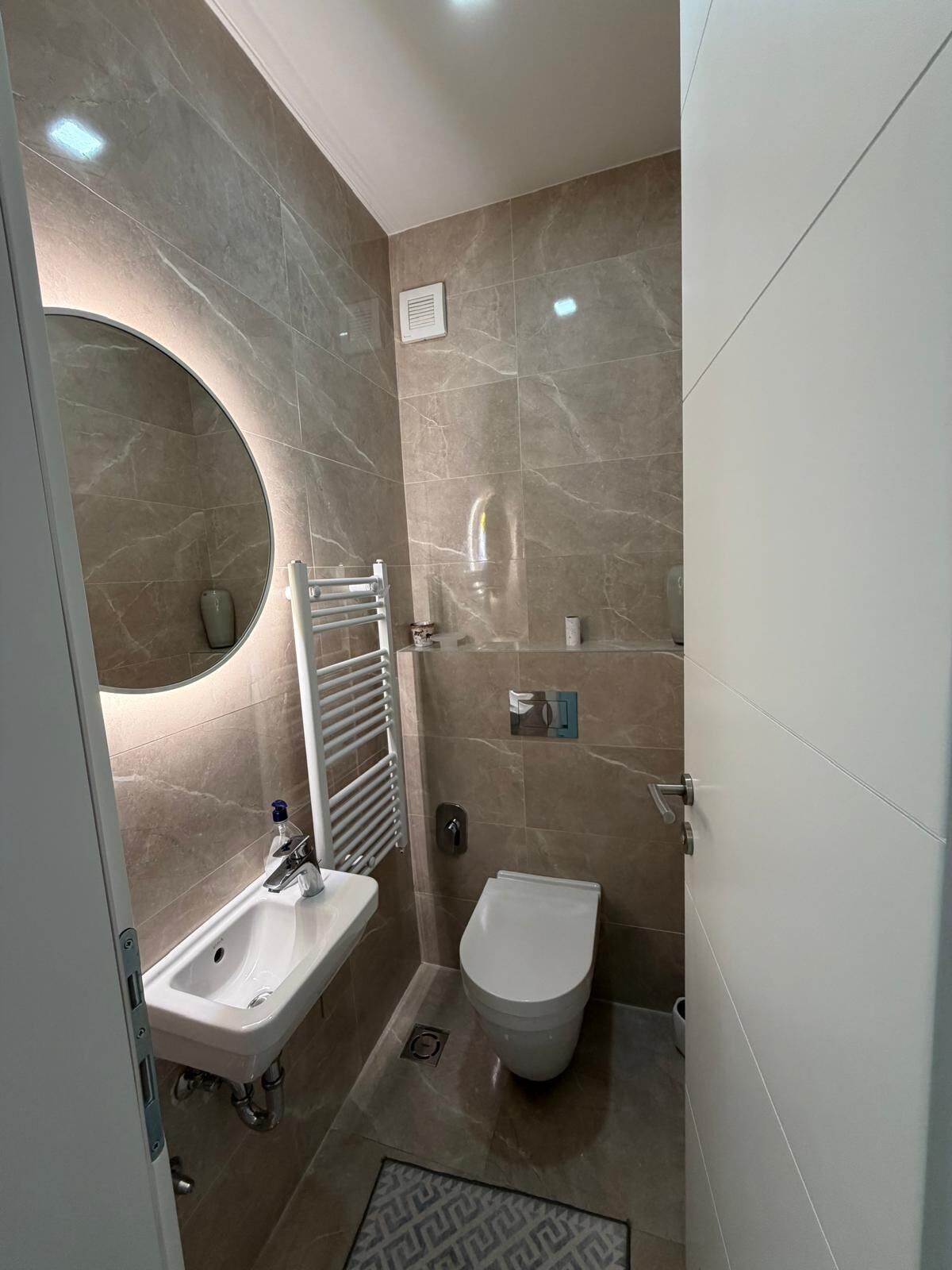 Een moderne badkamer met een wastafel, toilet en bidet, met marmeren wanden en een ronde spiegel.