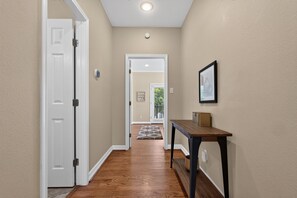 Un couloir avec un plancher en bois, une porte blanche, une petite table sur laquelle se trouve une boîte, et un tableau encadré accroché au mur.