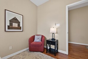 A room with a red chair, a small table with a lamp, a framed picture on the wall, and a wooden floor. - Central South Austin, Fenced Yard + Pets, Fire pit (Austin)