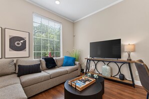 Ein Wohnzimmer mit einer Couch, einem Couchtisch mit Zeitschriften, einem Fernseher auf einem Schrank und einem Fenster mit Jalousien.