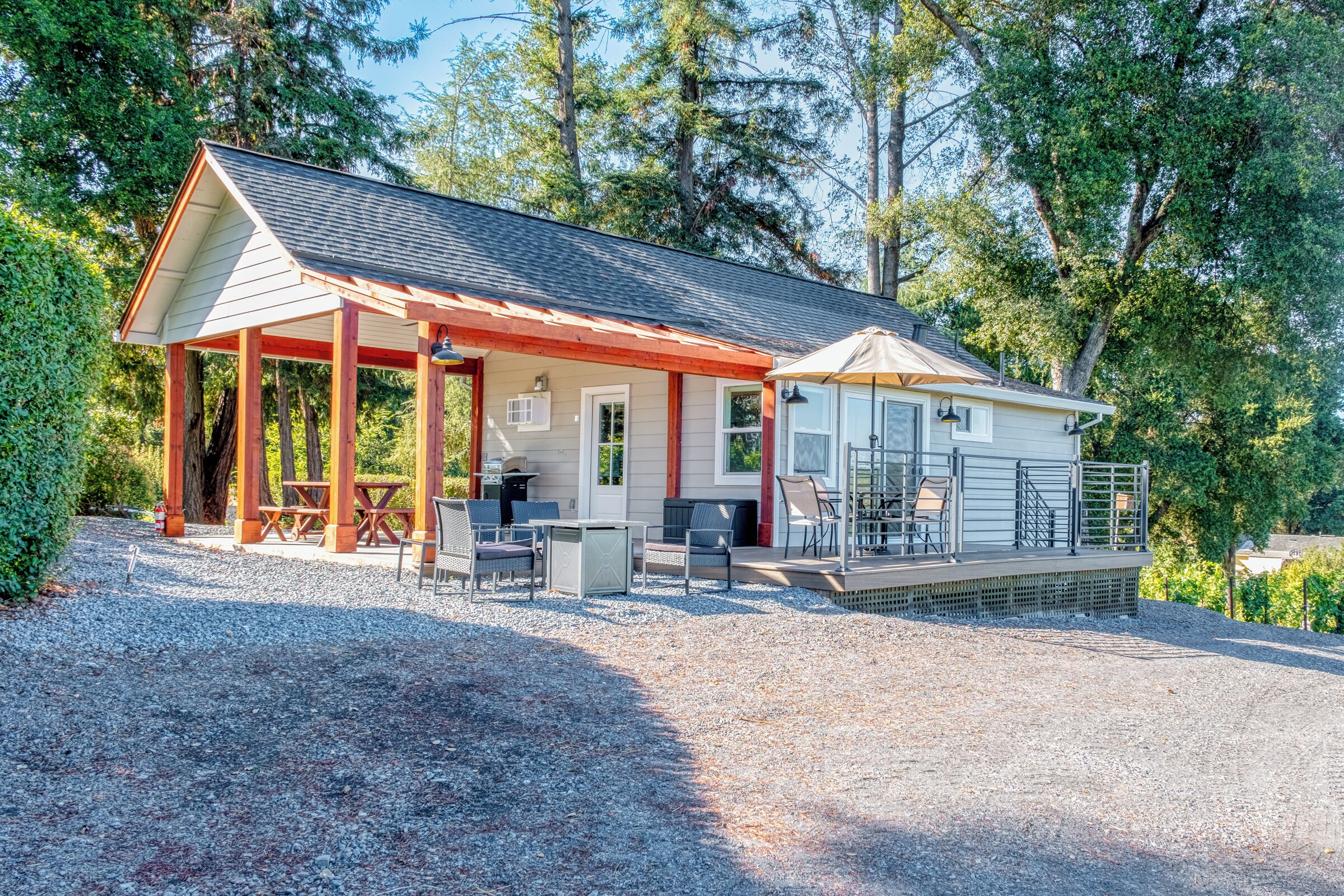 A covered outdoor area with a dining table and chairs, a small building with a balcony, and a gravel driveway.