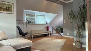 Un salon moderne avec un canapé, un fauteuil, un plancher en bois, un radiateur, une fenêtre avec des stores, une plante suspendue et un tableau au mur.