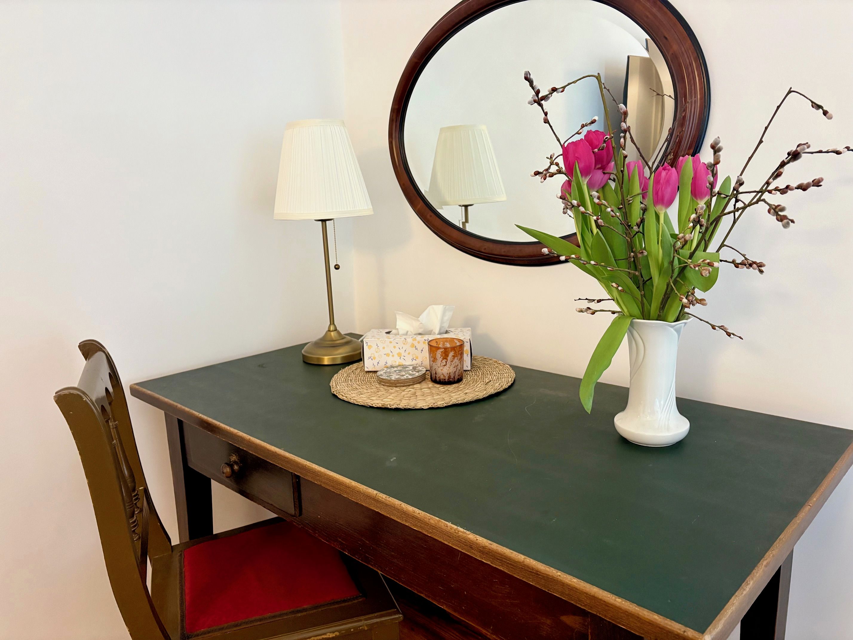 Un piccolo tavolo da trucco con uno specchio, una lampada, un vaso con fiori e un portasalviette.