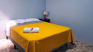 Una habitación pequeña con cama individual, colcha amarilla, almohadas blancas y una toalla blanca sobre la cama. Hay una mesita de noche con una lámpara junto a la cama.