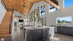 A spacious kitchen with dark cabinets, a large island, and a wooden staircase. - Waterfront 6-Acres w/ Pickleball, Hot Tub, Cold Plunge, Putting Green, Games! (Broken Bow)