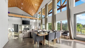 A spacious living area with a high ceiling, large windows, a fireplace, and a dining area with modern furniture. - Waterfront 6-Acres w/ Pickleball, Hot Tub, Cold Plunge, Putting Green, Games! (Broken Bow)