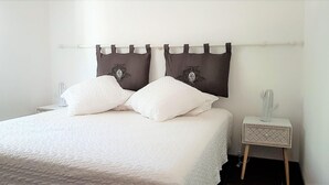 Ein ordentlich bezogenes Bett mit weißer Bettwäsche und zwei Zierkissen, ein Nachttisch mit Lampe und eine Wandvorhangstange.