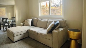 Un salon avec un canapé d’angle beige, une table à manger avec des chaises et une grande fenêtre donnant sur une terrasse en bois et des arbres.