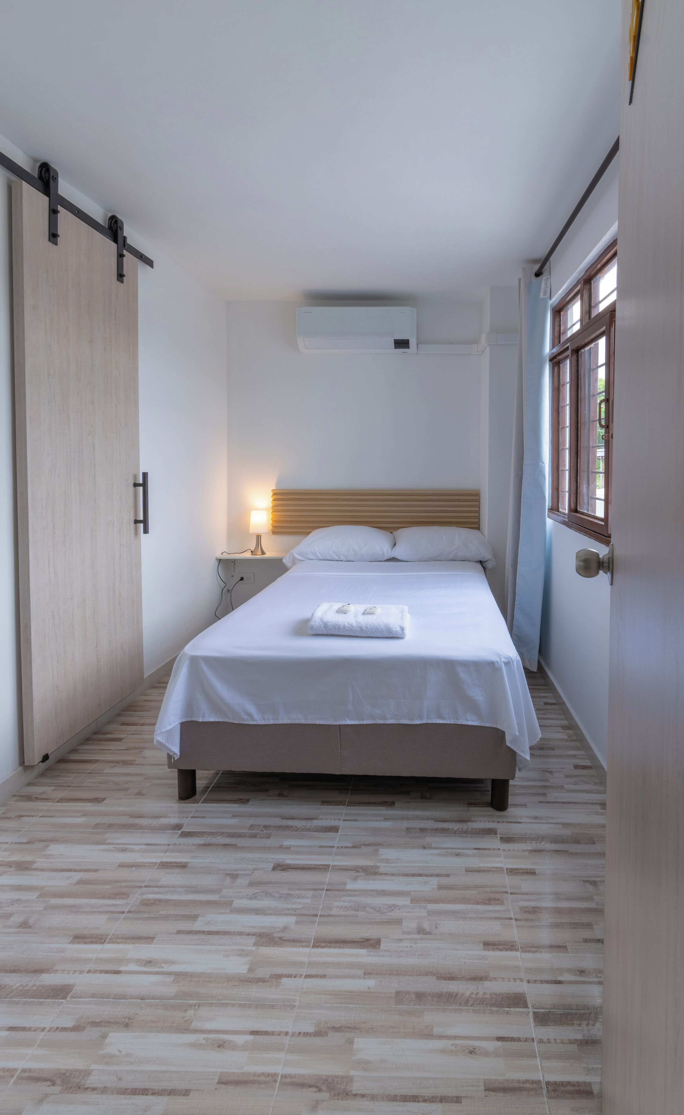 A bedroom with a bed, bedside table, lamp, and an air conditioning unit.
