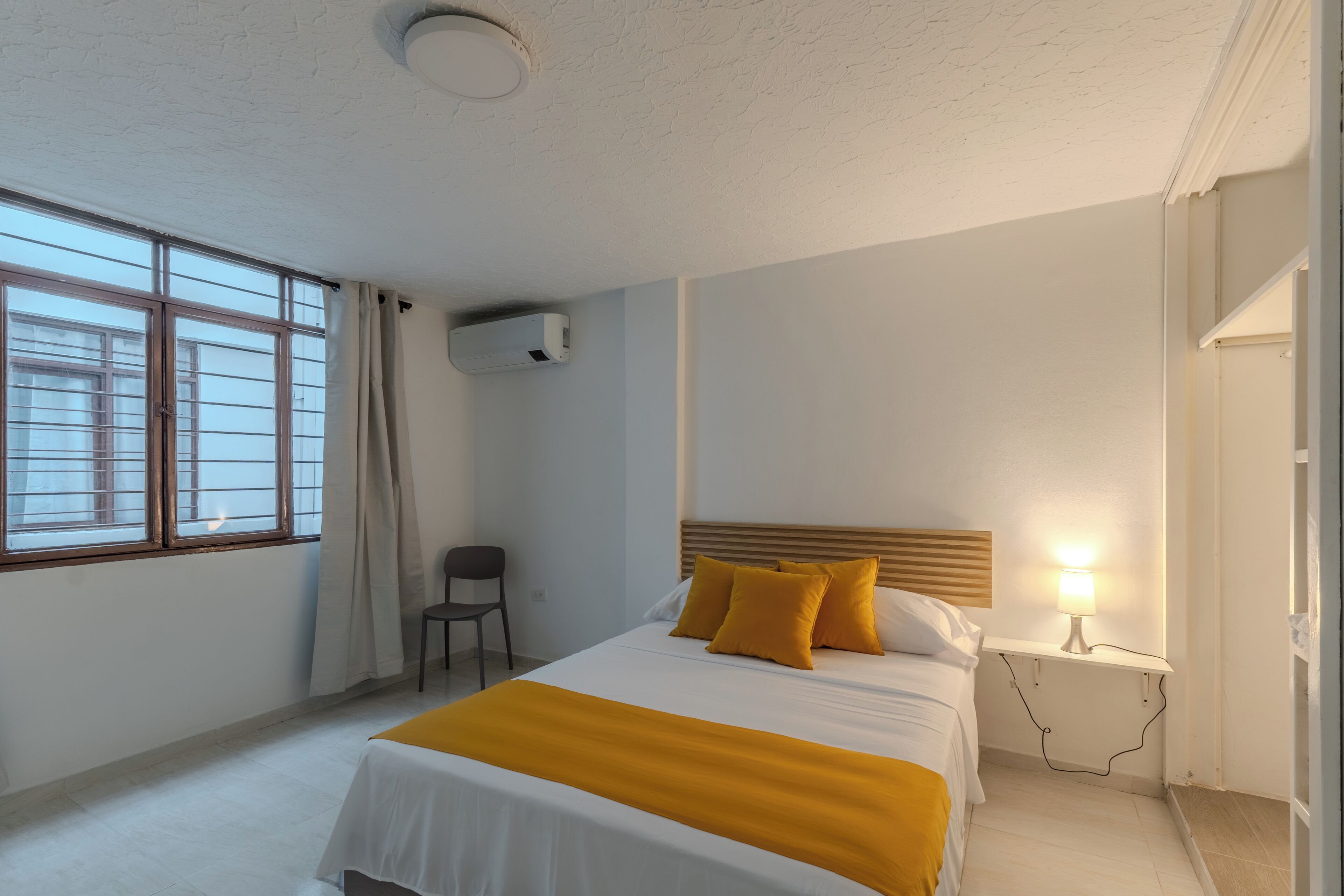Une chambre d’hôtel avec un lit, une chaise, une table de chevet avec une lampe et une fenêtre avec des rideaux.