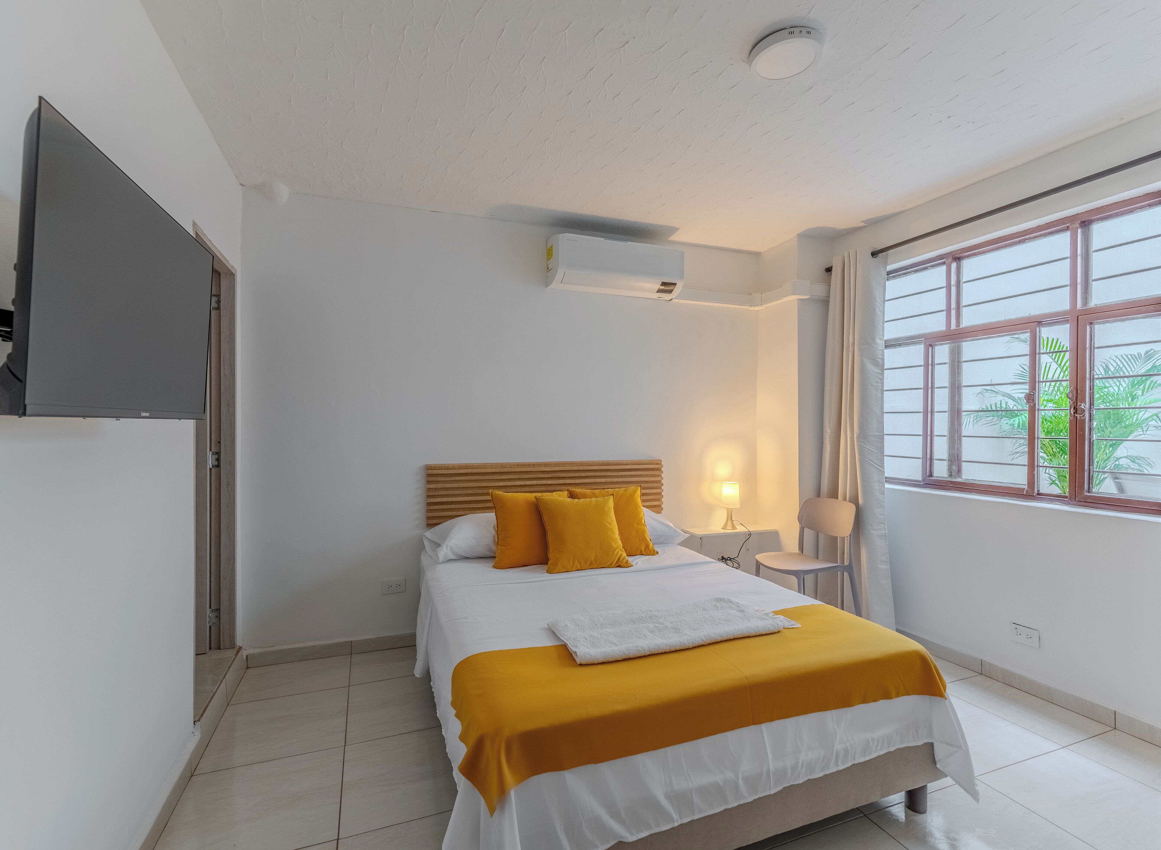 A hotel room with a bed, a bedside table with a lamp, a TV mounted on the wall, and a window with curtains.