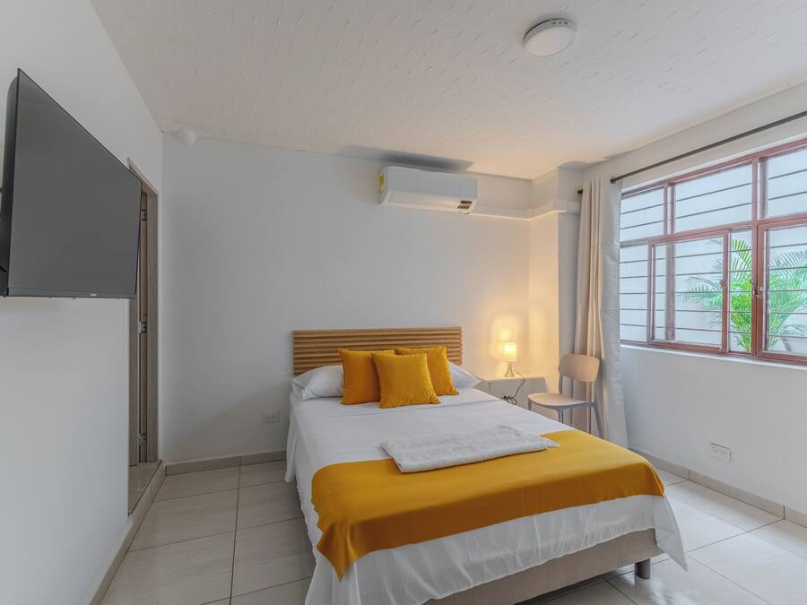 Habitación de hotel con una cama, una mesita de noche con lámpara, un televisor montado en la pared y una ventana con cortinas.