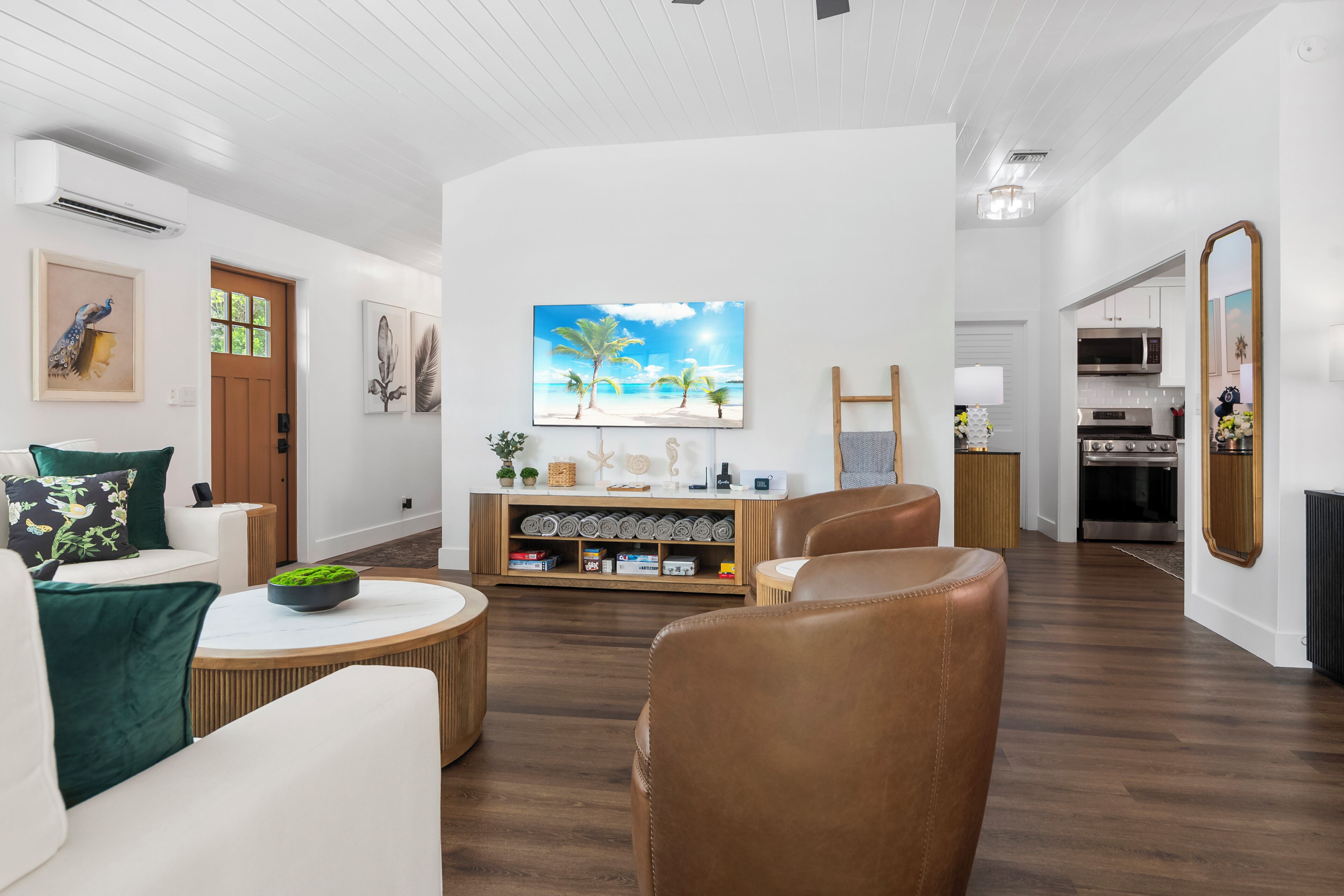 A modern living room with a wooden floor, white walls, and a large painting of a beach scene. There is a white sofa with green cushions, a brown leather chair, and a small round table with a bowl of green plants. A wooden cabinet with decorative items is against the wall.