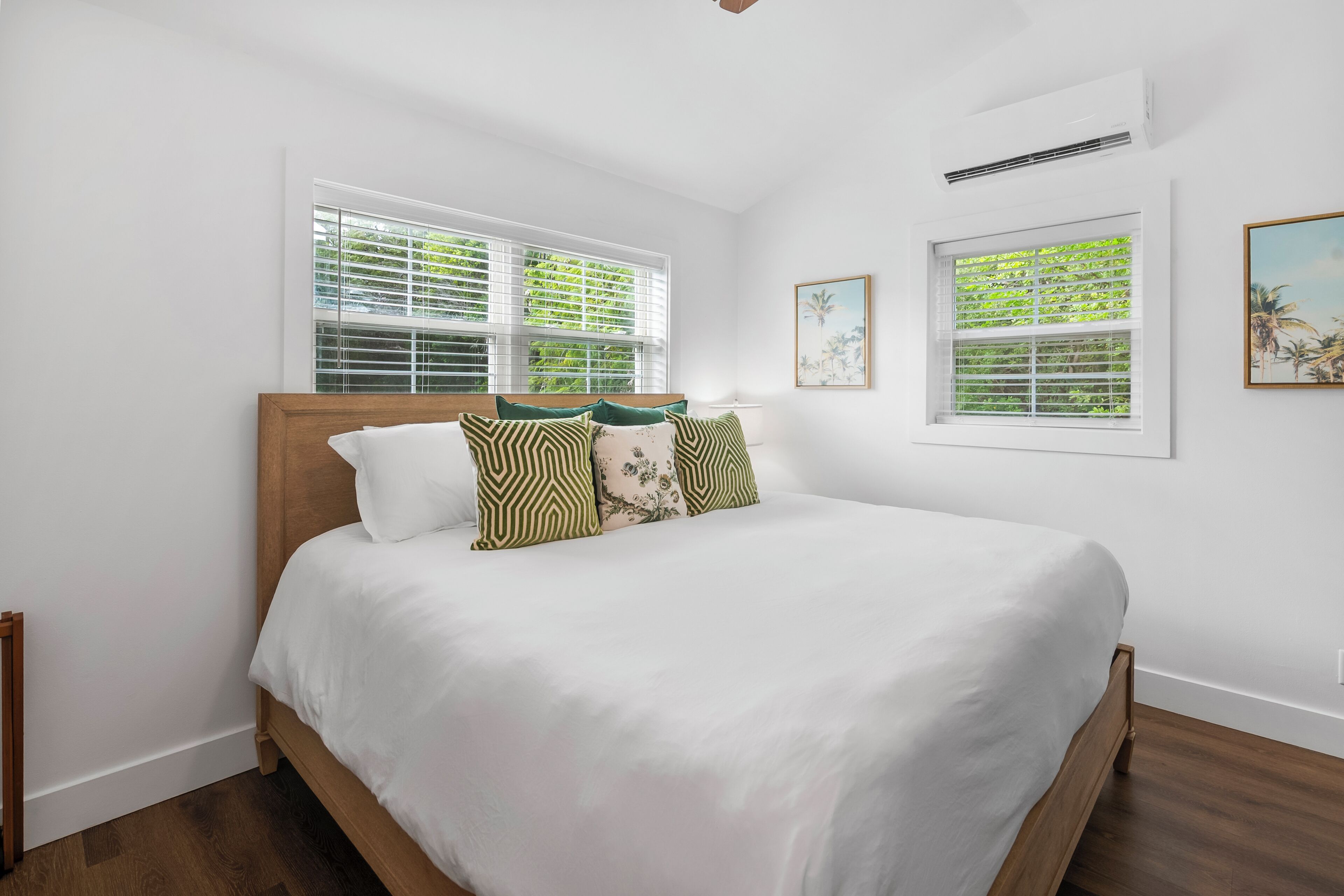 A bedroom with a large bed, two windows with blinds, a ceiling fan, and two framed pictures on the wall.