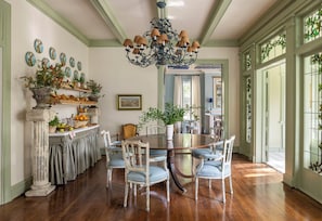 A dining room with a round table, chairs, and a chandelier. There are potted plants and a fruit basket on the sideboard. - The Marlene Inn (Houston)