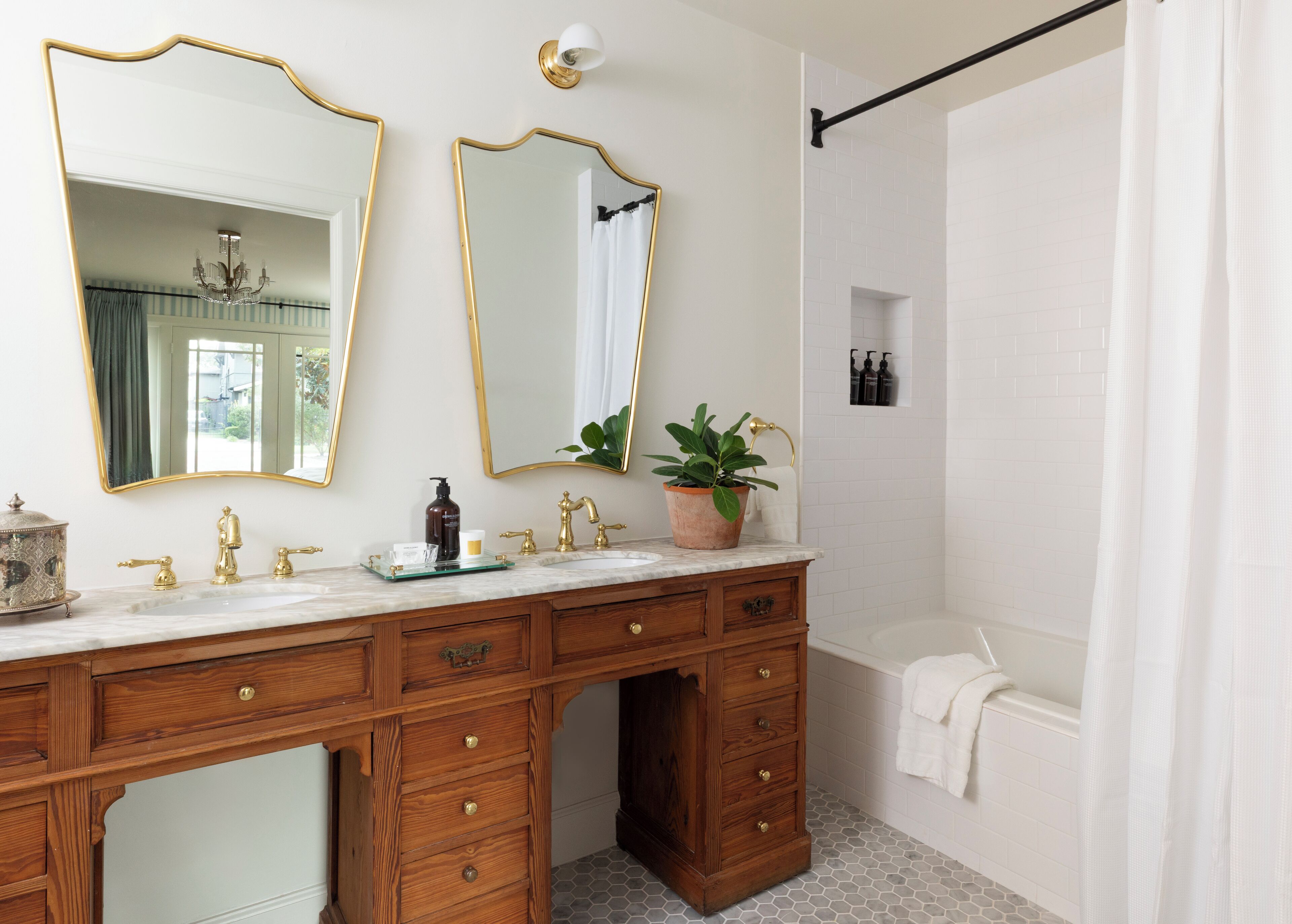 A bathroom with a wooden vanity, two gold-framed mirrors, a marble countertop, a potted plant, a shower, and a white tiled wall.