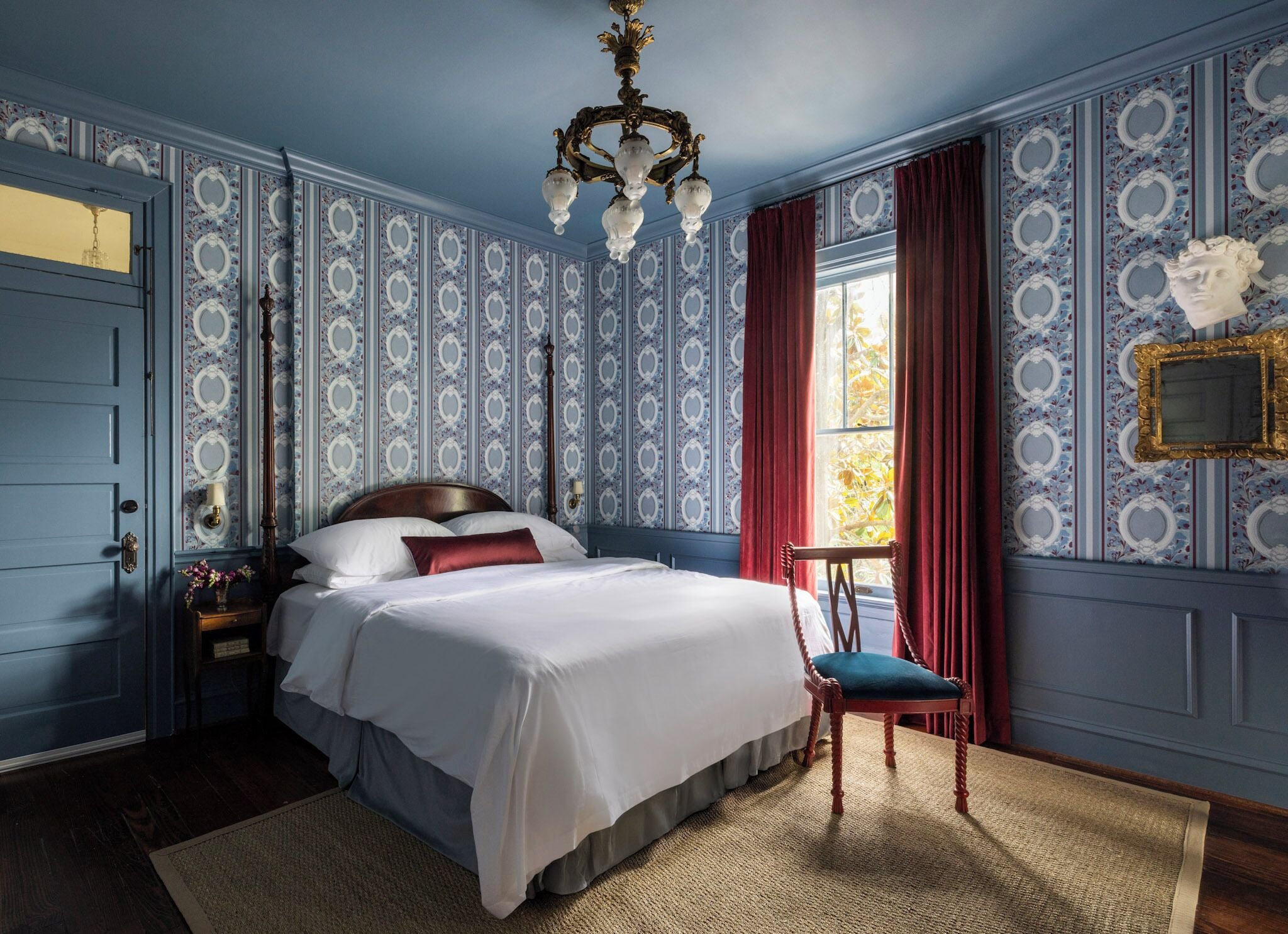 A bedroom with a large bed, a wooden chair, a chandelier, and patterned wallpaper.