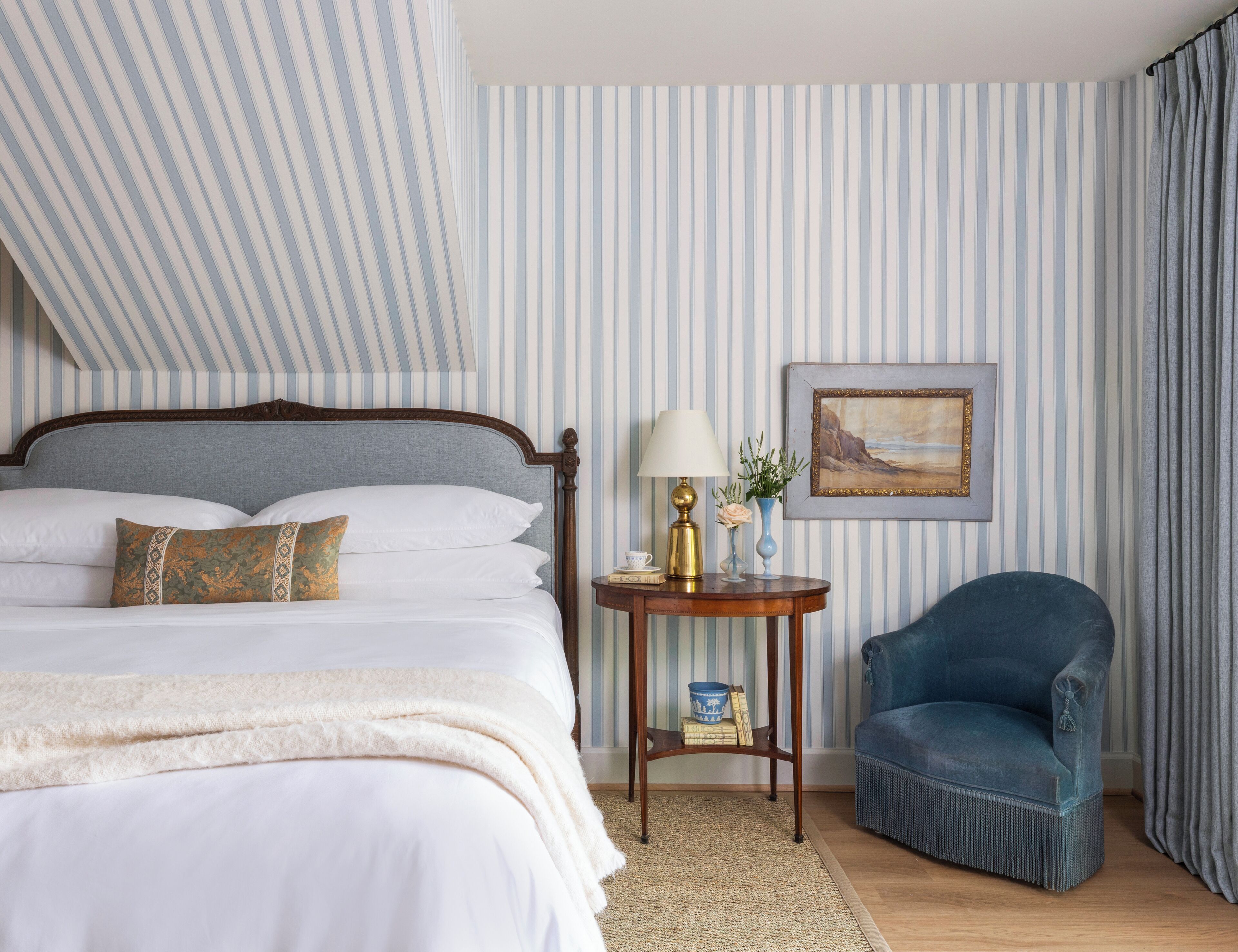 Una cama bien hecha con sábanas blancas y almohadones decorativos, una mesita de noche con lámpara y jarrón, un cuadro en la pared y un sillón azul.