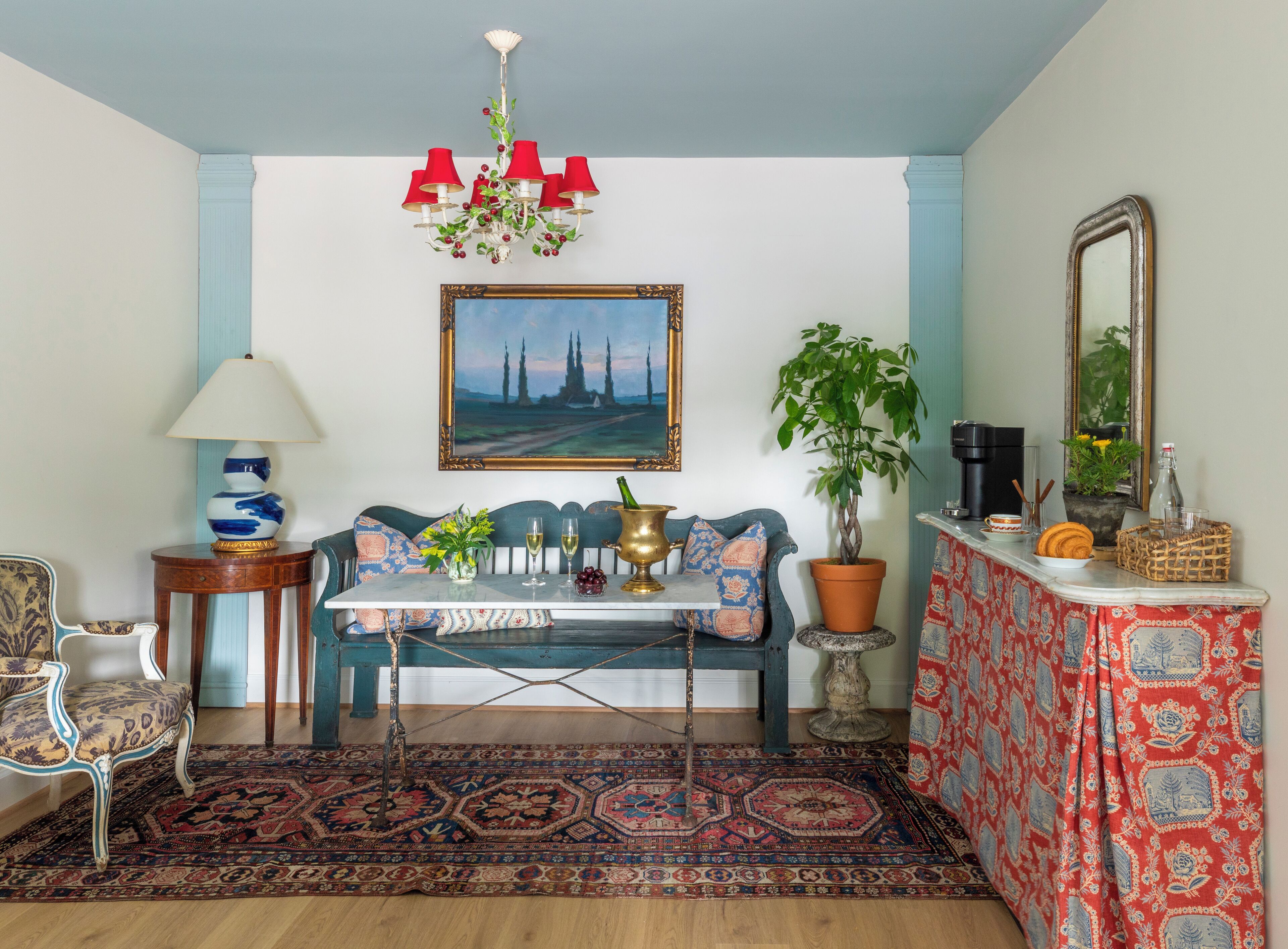 Ein Zimmer mit einem blauen Sofa, einem Tisch mit einer Vase und Weingläsern, einer Kandelaber, einem Gemälde, einer Topfpflanze und einem gemusterten Teppich.