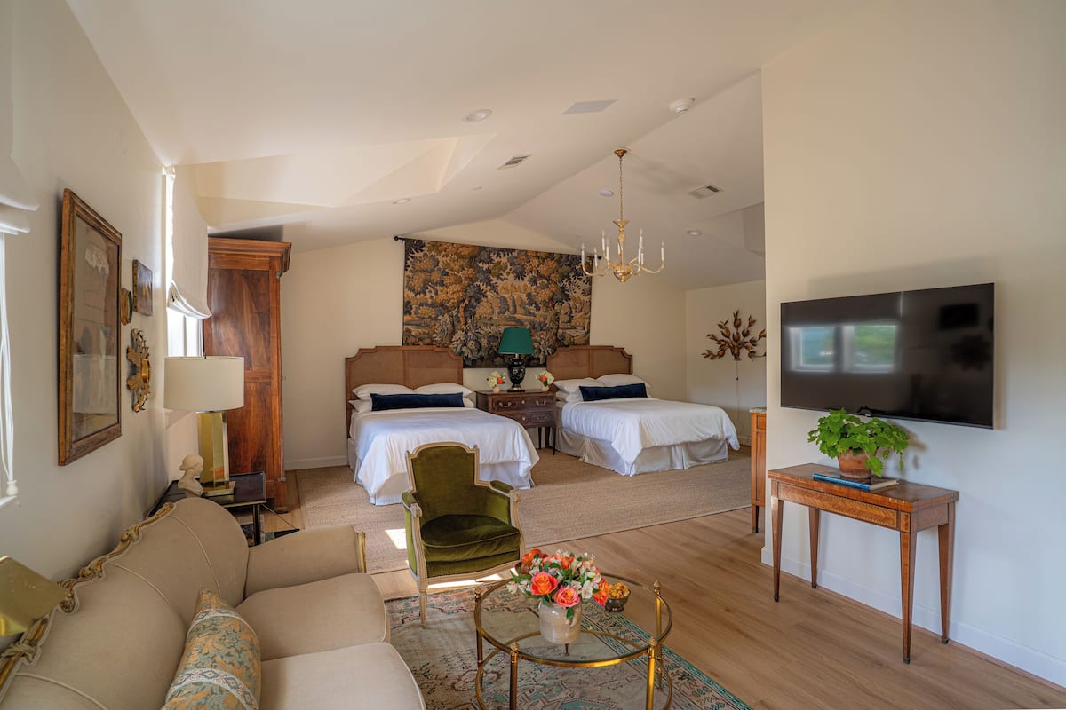 Una habitación amplia con una cama grande, un sillón verde, una mesa de centro con flores y un televisor en la pared.