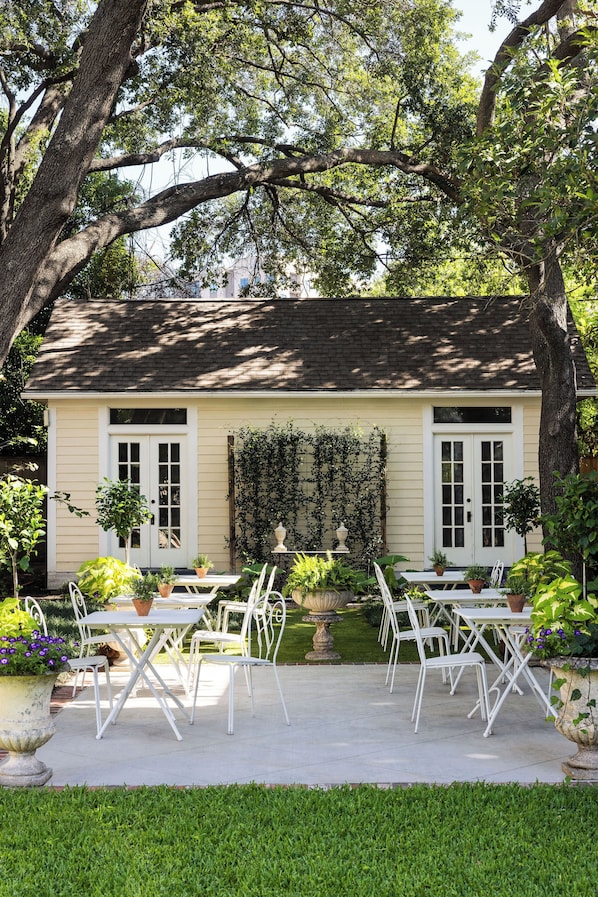 A house with a garden patio featuring white metal chairs and tables, surrounded by lush greenery and large trees. - The Marlene Inn (Houston)