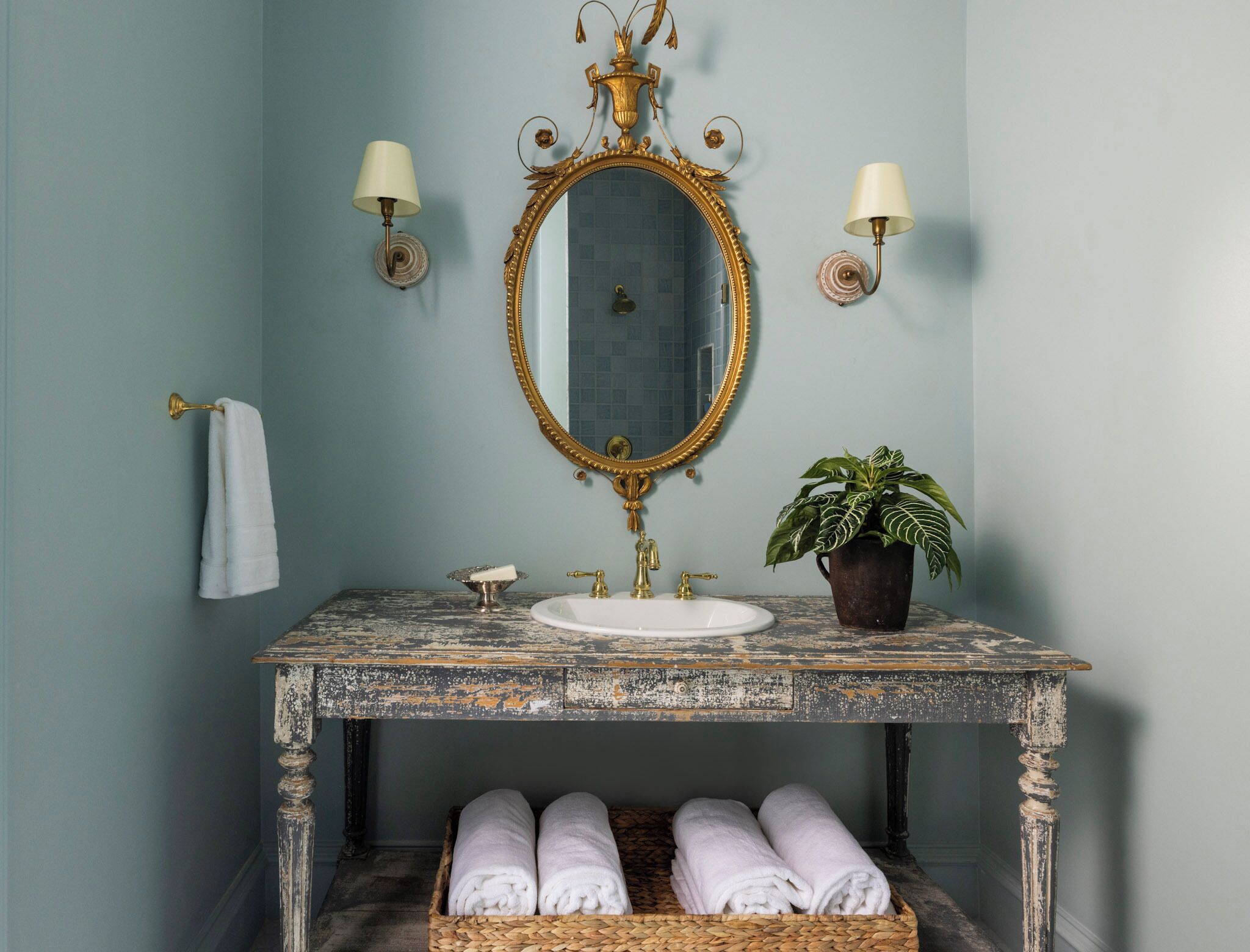 Une salle de bain au style rétro, avec un lavabo en marbre vieilli, un miroir encadré d'or, une plante en pot et un panier en osier contenant des serviettes.
