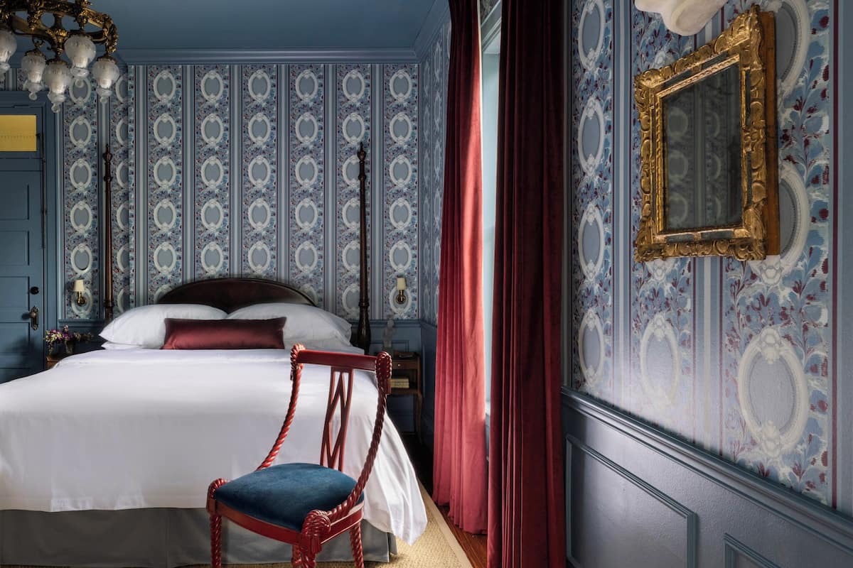 Un dormitorio con una cama grande, una silla de madera, una araña y papel tapiz estampado.