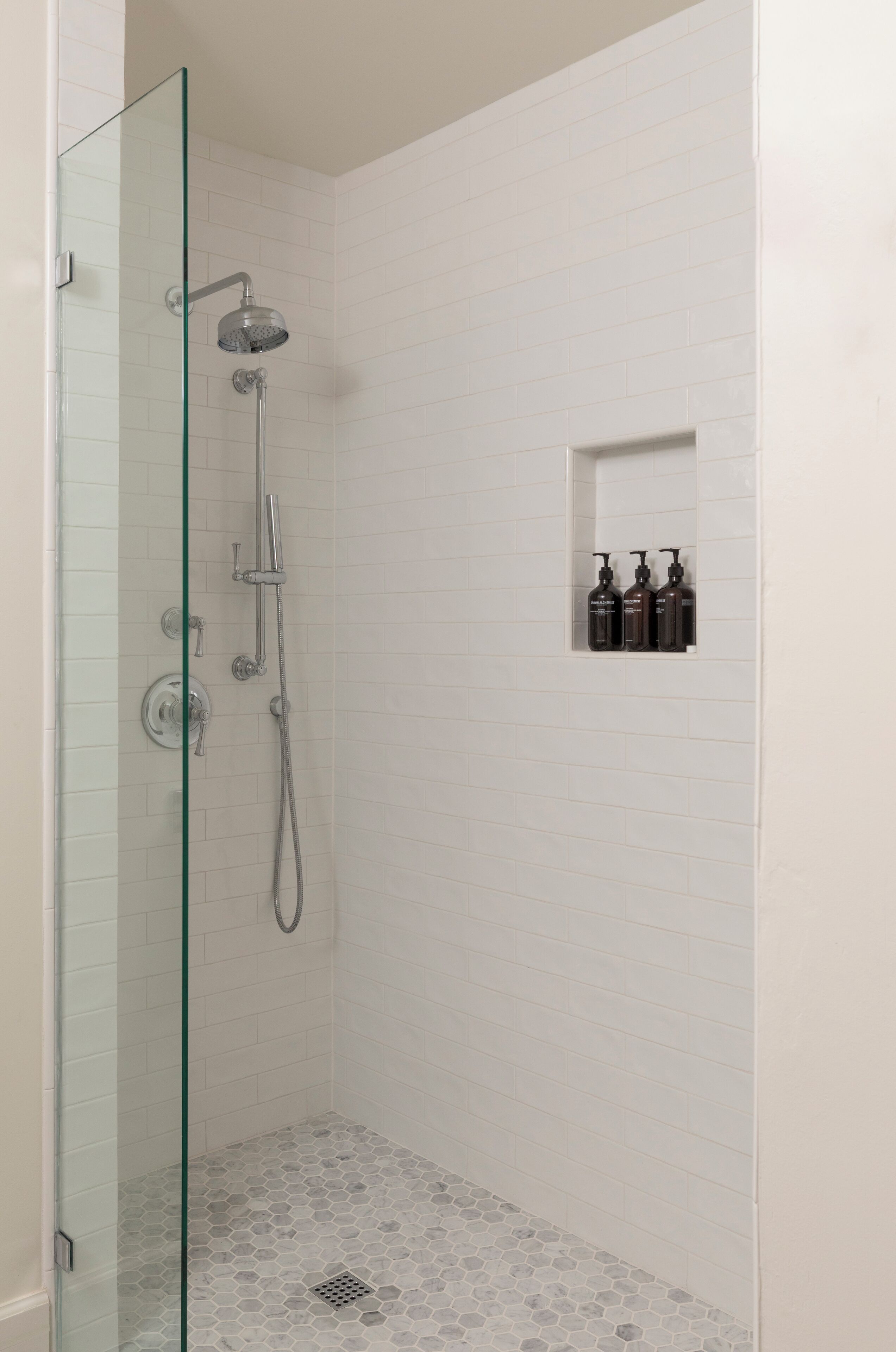 Une douche fermée par des parois de verre, avec un mur recouvert de carreaux blancs, une petite étagère pour les articles de toilette et un sol à motifs hexagonaux.