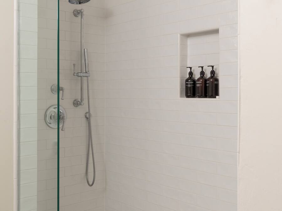 A glass-enclosed shower with a white tiled wall, a small shelf with toiletries, and a hexagonal patterned floor.