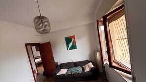 A room with a dark sofa, a white bed, a hanging light fixture, and a window with a view. - Casa Rina Family home apartment. Umbria / Tuscany. 2 minutes from the lake (Umbria)