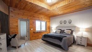 Ein Schlafzimmer mit Holzboden, einem Bett mit grauer Bettwäsche, einem Nachttisch mit Lampe, einem Sessel und einem Fenster.