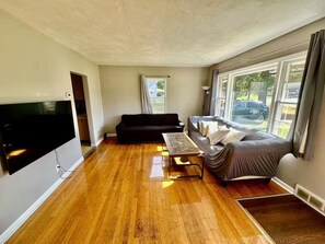 A spacious living room with hardwood flooring, a flat-screen TV mounted on the wall, a black sofa, a grey sofa, and a small coffee table.