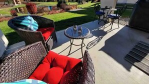 An outdoor patio with wicker furniture and a glass table, overlooking a landscaped garden and mountains. - Enjoy our beautiful two-bedroom condo over Christmas break. (Leavenworth)