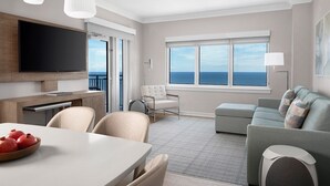 A modern living room with a flat-screen TV, a dining table with a bowl of pomegranates, a grey sofa, and a view of the ocean through large windows. - 2-bedroom condo steps to beach! Marriott Beach Place Towers (Fort Lauderdale)
