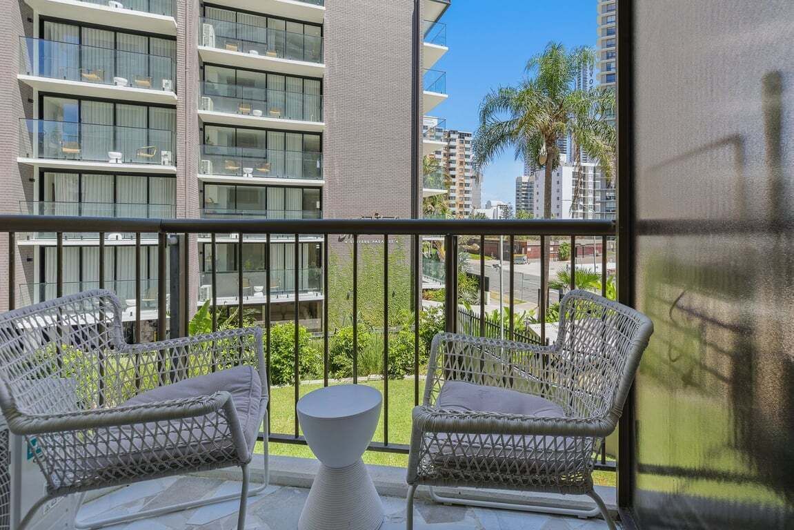 Un balcón con sillas de mimbre y una mesita, con vistas a un paisaje urbano con edificios modernos y palmeras.