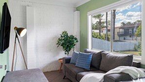 Une pièce avec un mur en briques blanches, un canapé gris avec des coussins bleus, un plaid blanc, une lampe sur pied et une fenêtre donnant sur des maisons.