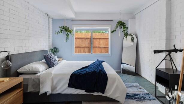 Une chambre à coucher avec un lit, une table de chevet, une plante suspendue, un télescope et une fenêtre donnant sur une clôture en bois.