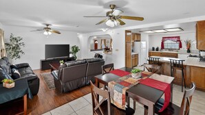 Ein modernes Wohnzimmer mit Sofa, Fernseher und Deckenventilatoren. Ein Essbereich mit Tisch und Stühlen ist ebenfalls zu sehen.
