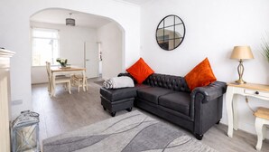 Een moderne woonkamer met een zwarte leren bank, twee oranje kussens, een ronde spiegel en een lamp op een houten bijzettafeltje.