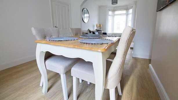 Een eetgedeelte met een witte tafel en stoelen, een spiegel aan de muur, en een raam met natuurlijk licht.