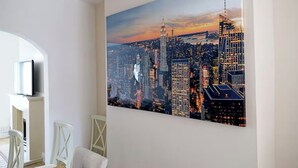 Een stadsgezichtschilderij aan een muur, een witte stoel en een witte tafel met een flatscreen televisie.