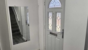 Een gang met een witte deur, voorzien van een glas-in-loodraam, een kleine spiegel aan de muur en een donker houten vloer.