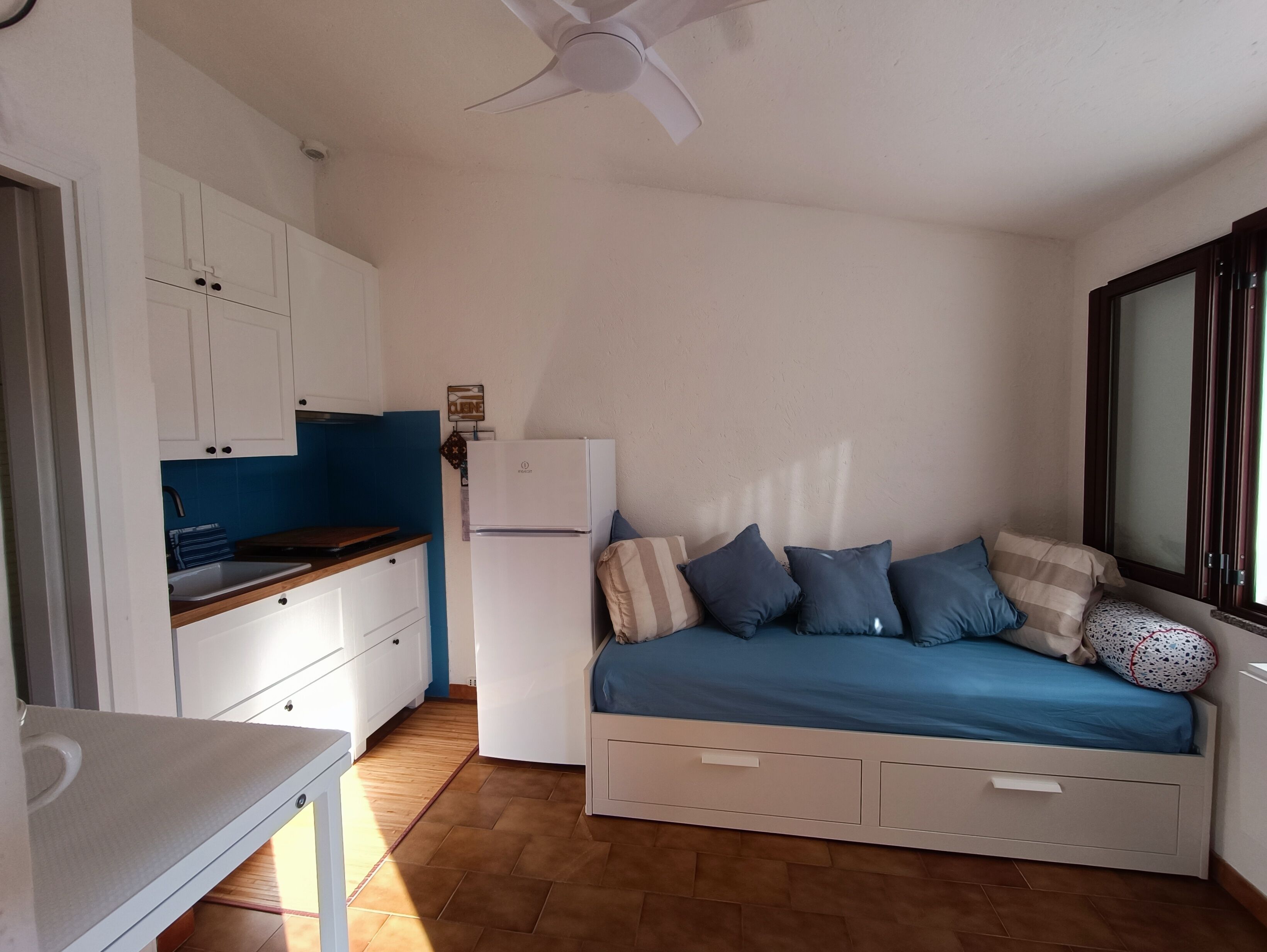 Ein kleines Zimmer mit Küchenzeile, einem Bett mit blauer Bettwäsche und einem Deckenventilator.