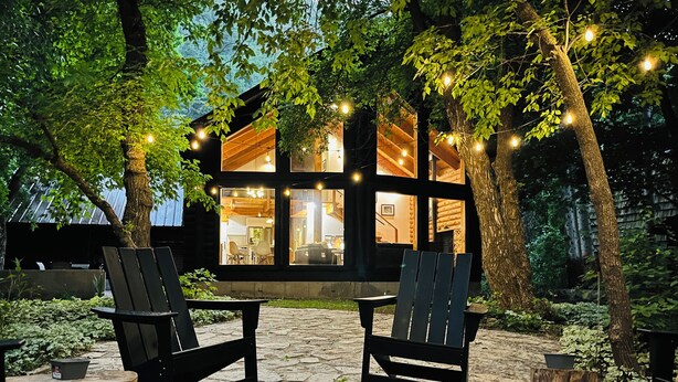 Un espace extérieur accueillant, aménagé avec deux fauteuils Adirondack et un foyer en pierre, entouré d’arbres et d’un chalet en bois illuminé de guirlandes lumineuses.