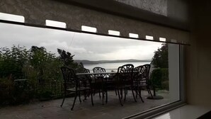 Udsigt fra et vindue over en patio med et bord og stole, et vandareal og fjerne bakker.
