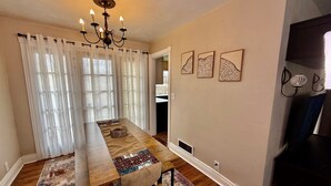 A dining area with a long wooden table, white curtains, and a chandelier. There are three wall-mounted artwork pieces and a wall-mounted lamp. - Garden Cottage | Close to SLC (Midvale)