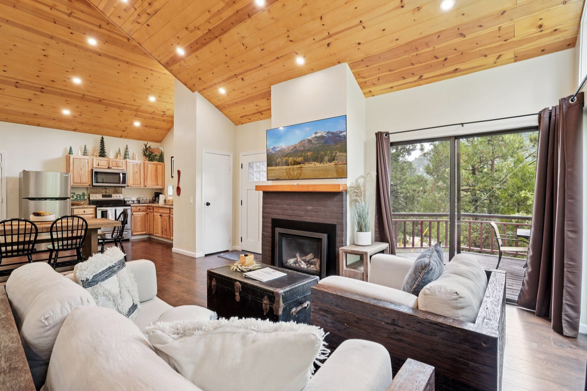 A cozy living room with a fireplace, wooden ceiling, and a large window overlooking a deck.