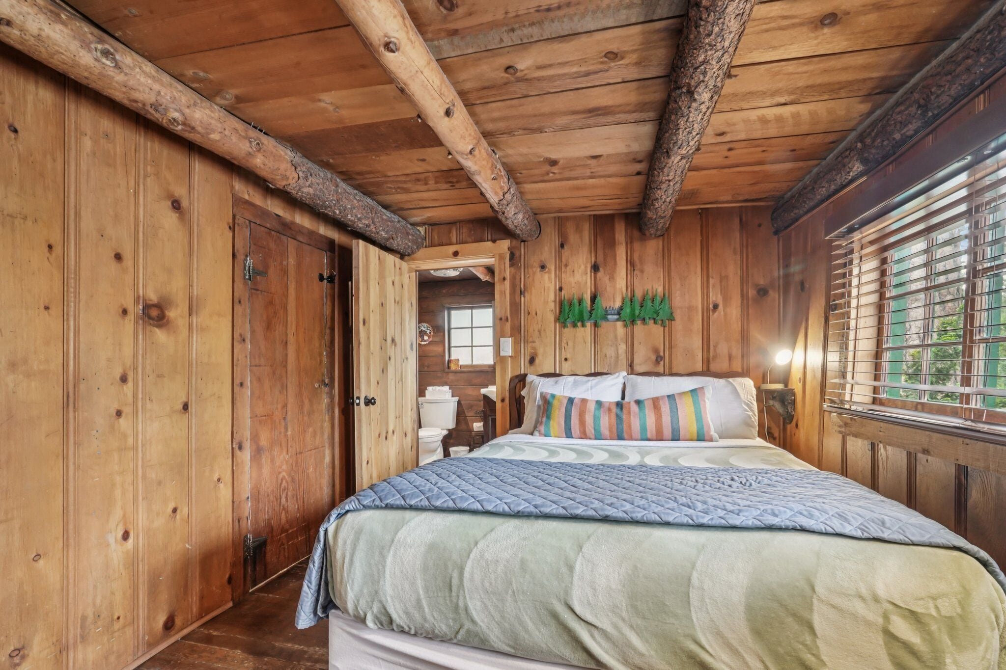 A wooden cabin bedroom with a bed, wooden walls, and a window with blinds.