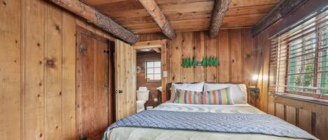 A wooden cabin bedroom with a bed, wooden walls, and a window with blinds.