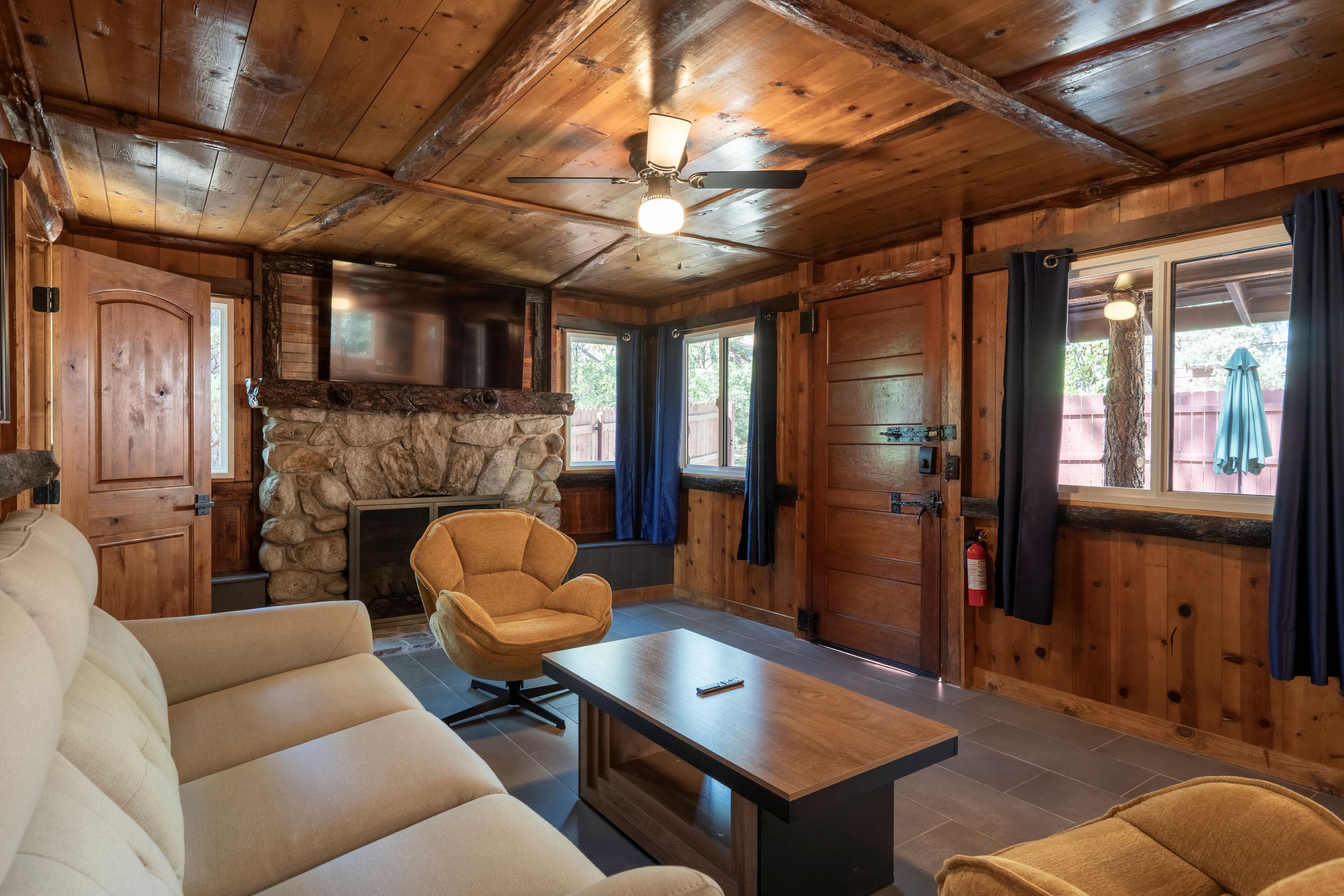 A cozy cabin interior with a stone fireplace, wooden walls, and a ceiling fan.